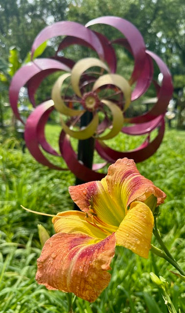 Metallic windmill sculpture with a pink and yellow flower in a garden setting